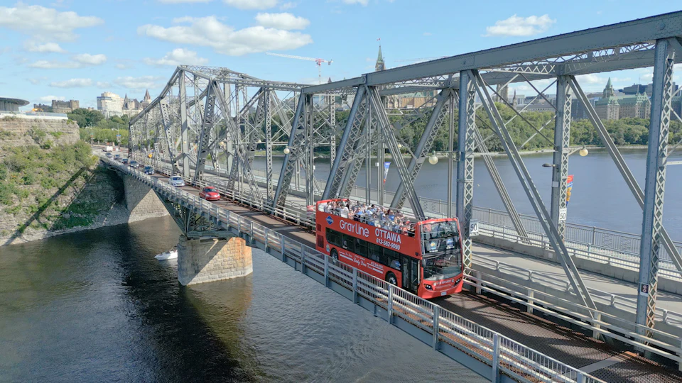 A red double-decker tour bus crossing a steel bridge over a river, with trees and buildings visible in the background.