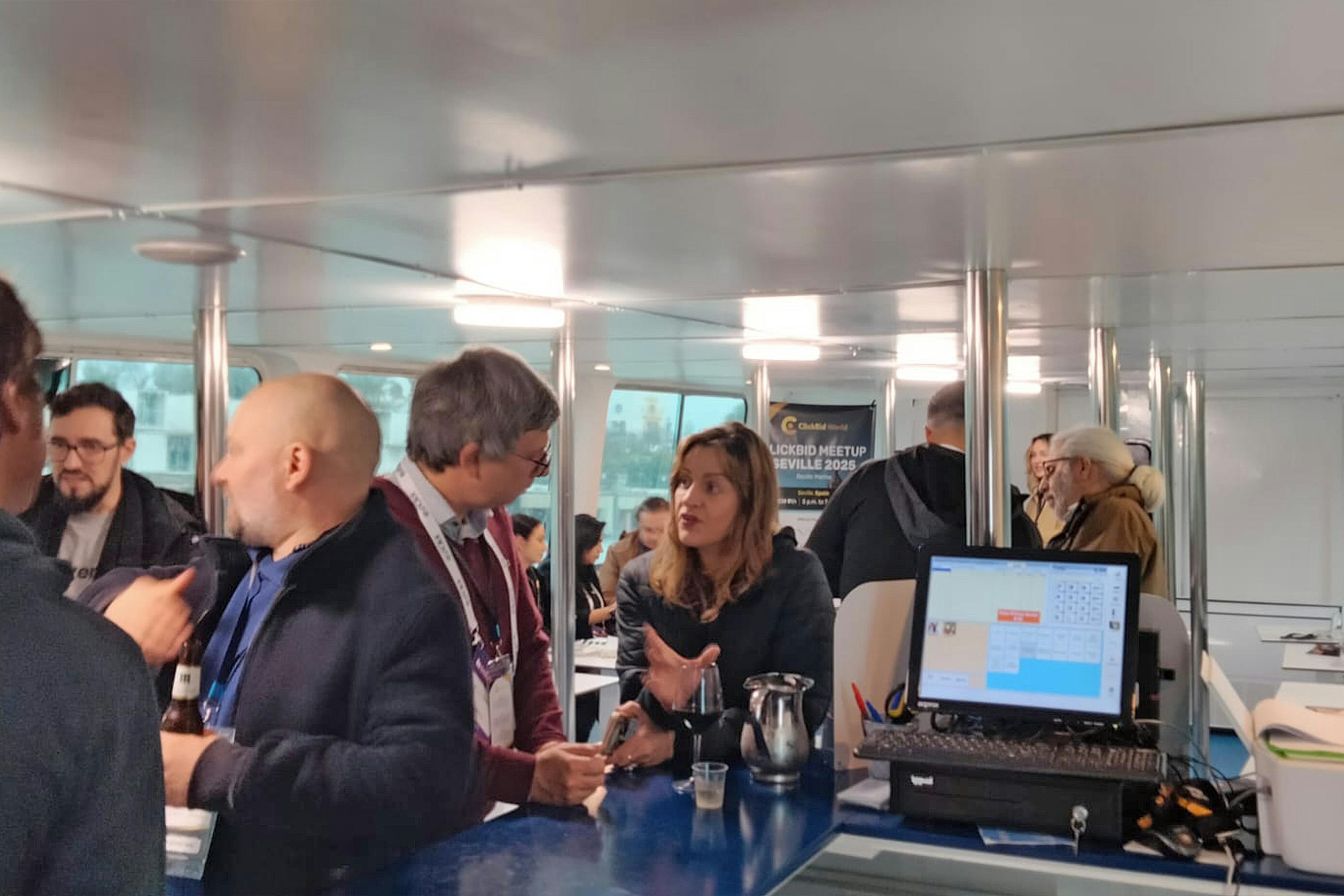 Nawiązywanie kontaktów i rozmowa na spotkaniu w pokoju z dużymi oknami. Na banerze jest napisane „CCd Meetup Sewilla 2023”.