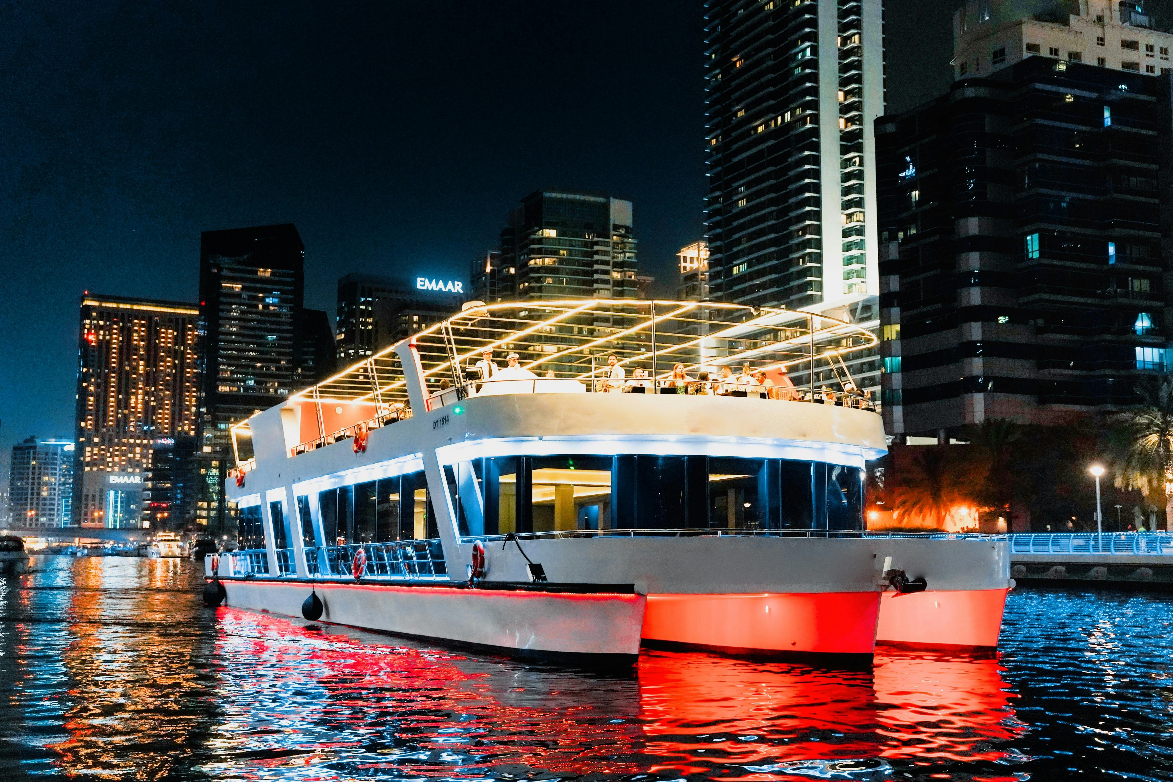 Moderní loď s osvětlenou horní palubou se v noci vznáší v městské řece s vysokými budovami v pozadí.