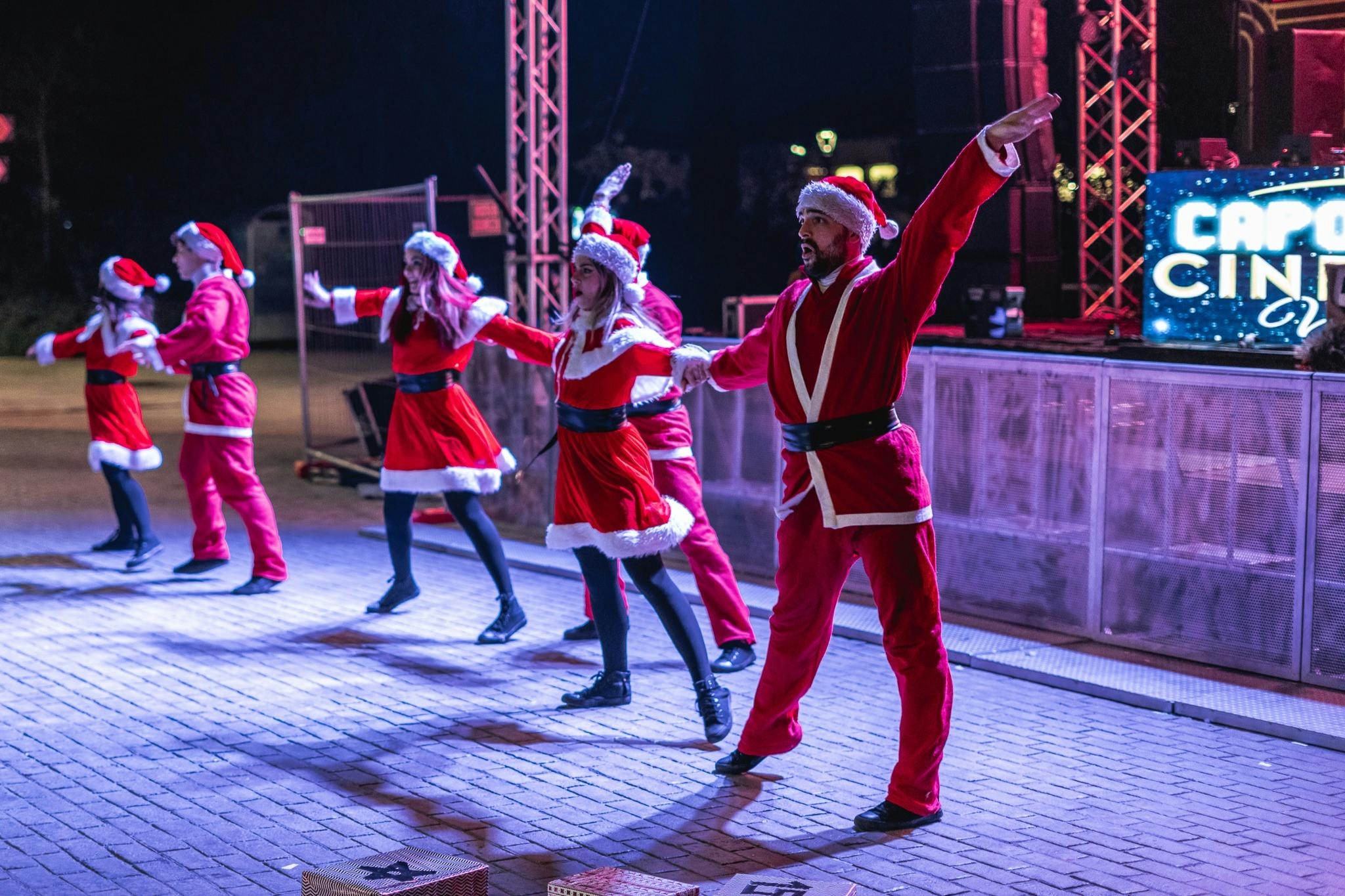 People dressed as Santa Claus perform a choreographed dance on a stage at night.
