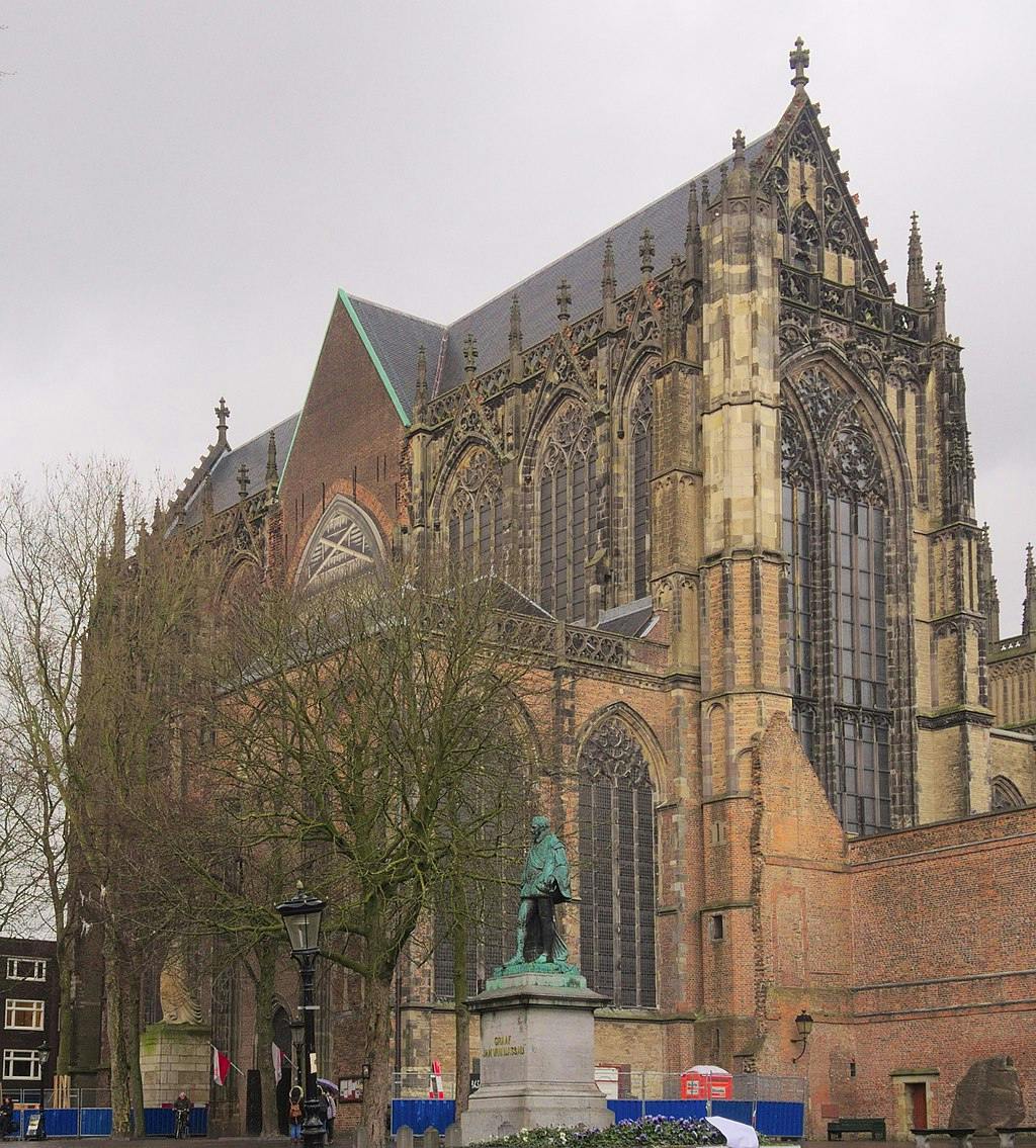 Церковь в готическом стиле с большими витражами, замысловатыми деталями и бронзовой статуей спереди, расположенной под серым небом.