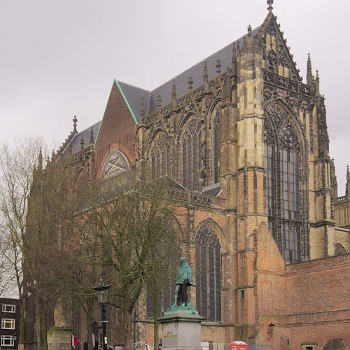Kyrka i gotisk stil med stora glasmålningar, intrikata detaljer och en bronsstaty framför, under en grå himmel.