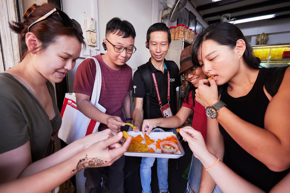 Four people share a tray of bright orange and yellow food. One person wears a hat and another has a bag. They appear to be enjoying the food together.