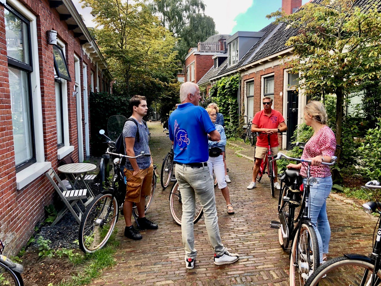 A group of people standing with bicycles on a narrow, cobblestone street lined with brick houses and greenery.