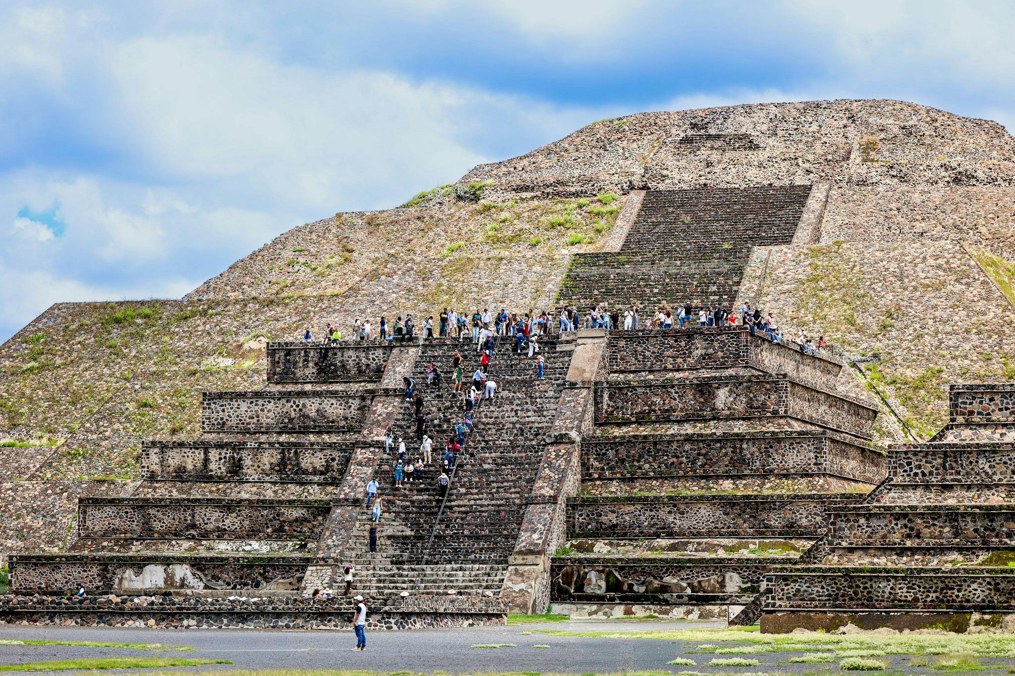 A pyramid-like stone structure with people ascending its steps, set against a partly cloudy sky, and a grassy foreground.