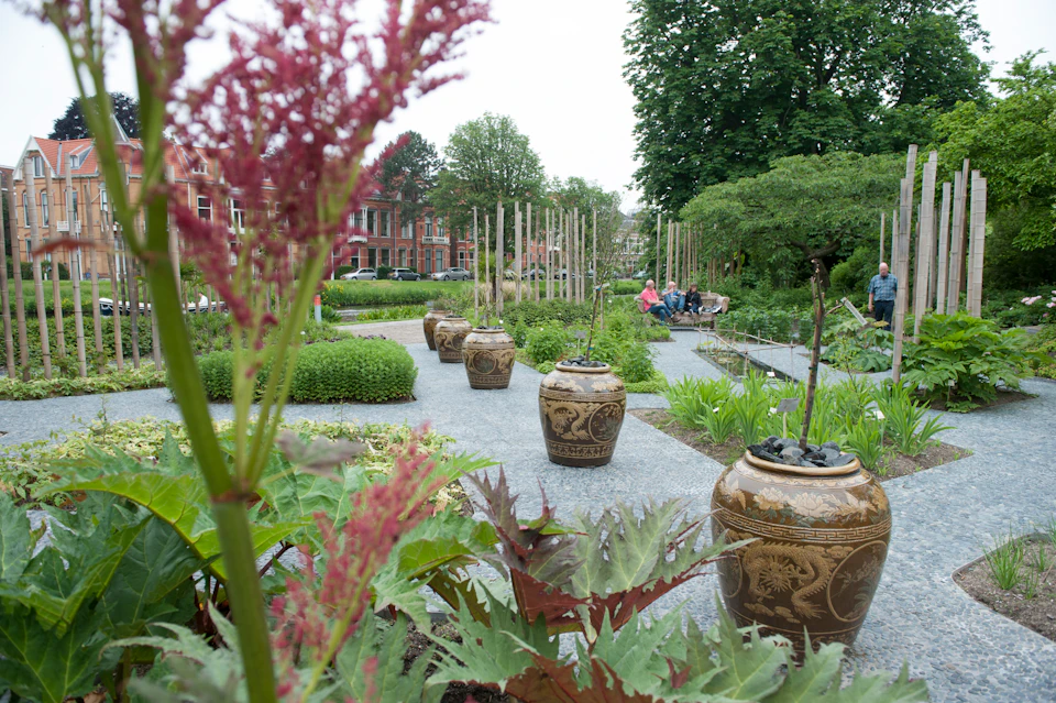 A scenic garden with ornamental pots, lush greenery, and people sitting on benches. Red brick buildings are visible in the background.
