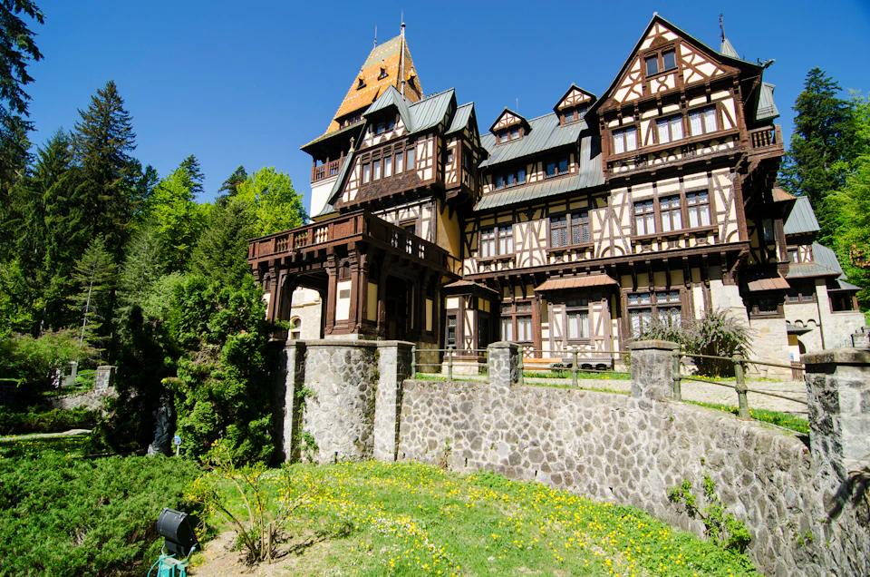 A large, ornate timber and stone building with balconies, surrounded by lush greenery on a clear, sunny day.