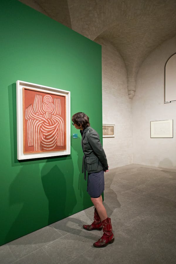 Ένα άτομο εξετάζει αφηρημένα έργα τέχνης σε έναν πράσινο τοίχο σε έναν μινιμαλιστικό χώρο γκαλερί με ψηλά ταβάνια και άλλη πλαισιωμένη τέχνη.