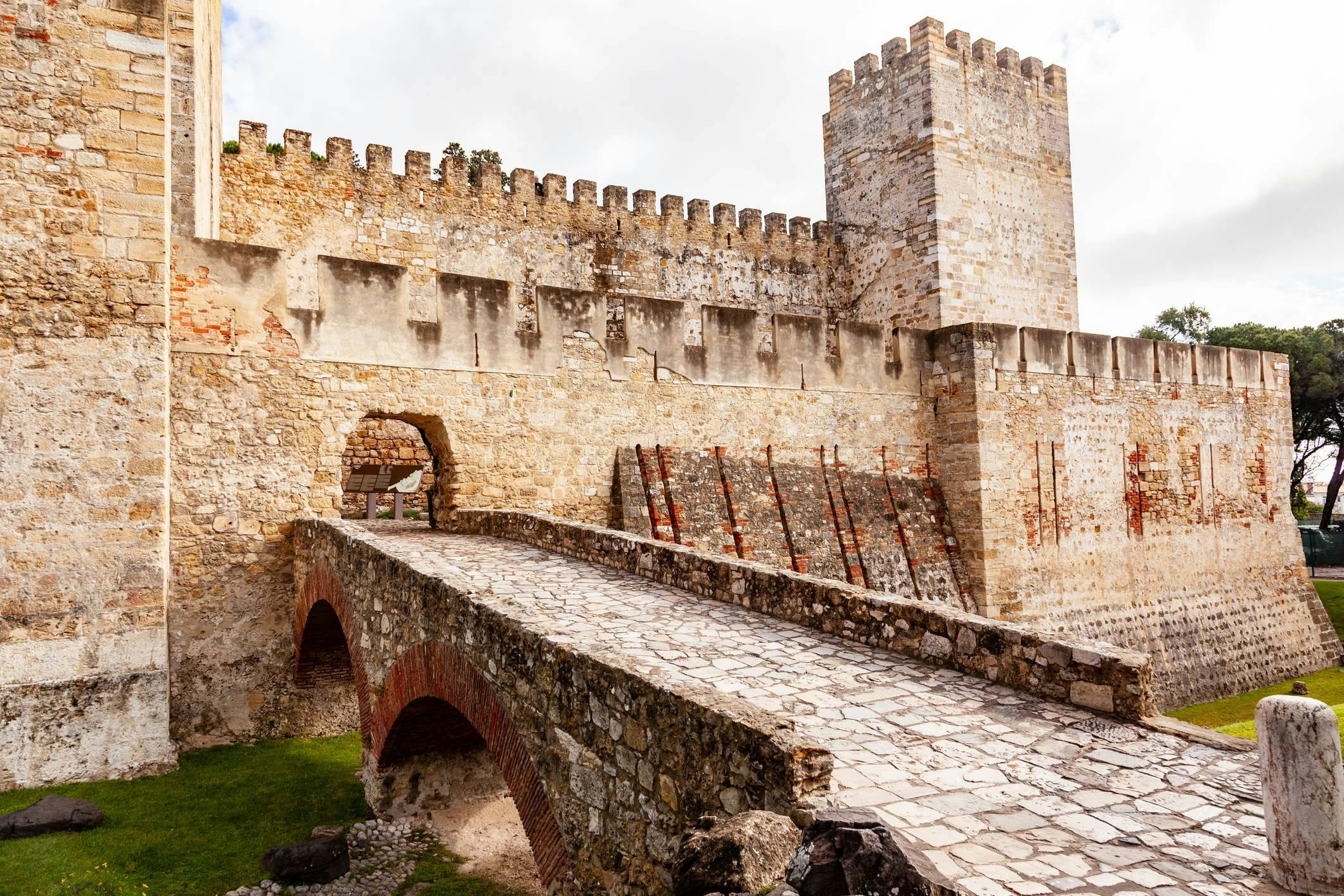 Stone bridge leading to a historic fortress with crenelated walls and a tall tower under a partly cloudy sky.