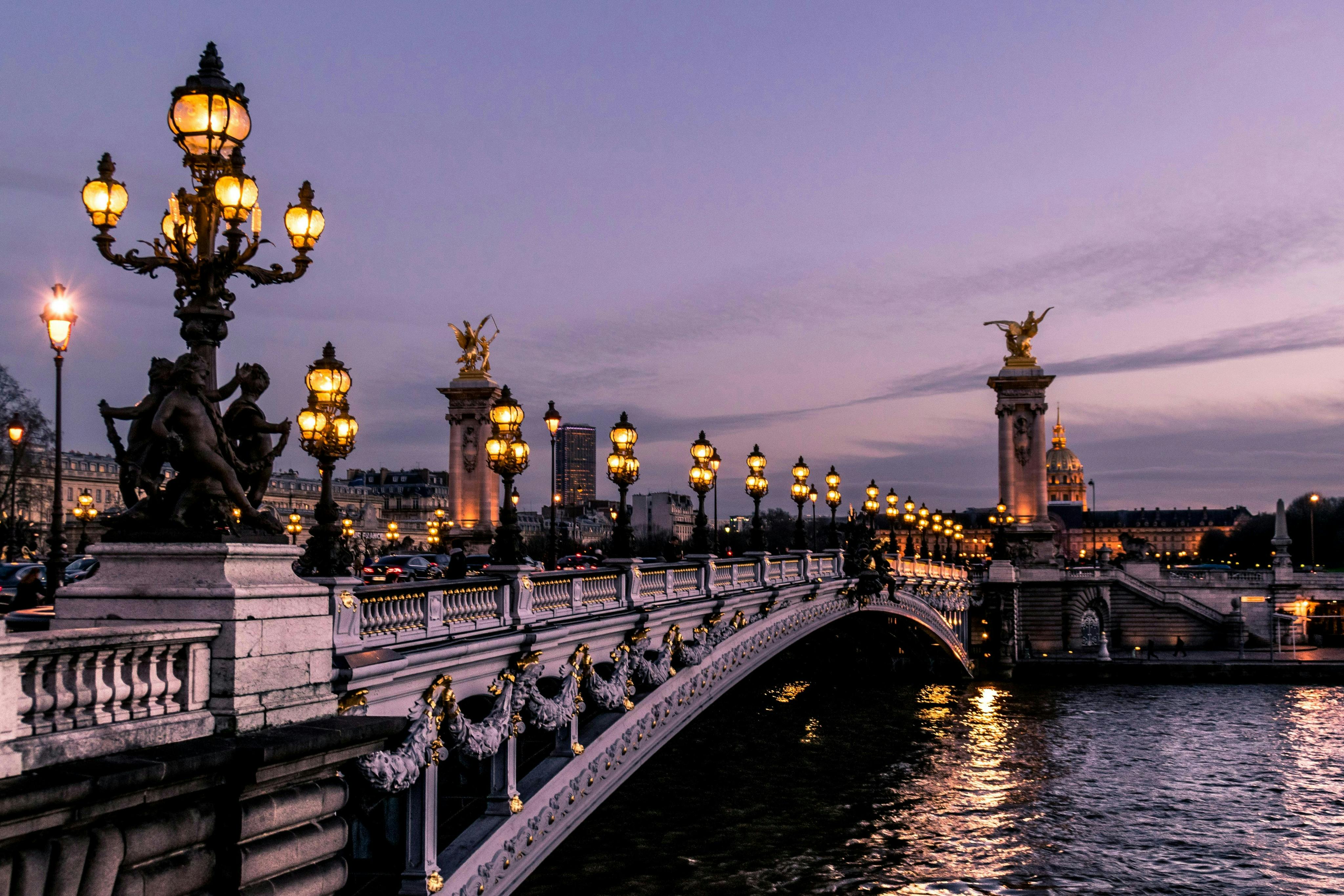 A bridge adorned with ornate streetlights and sculptures, illuminated at dusk with a cityscape in the background.