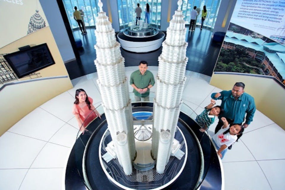 Dos personas de pie cerca de una detallada maqueta de las Torres Petronas expuesta en una moderna zona de exposición interior con grandes ventanales.