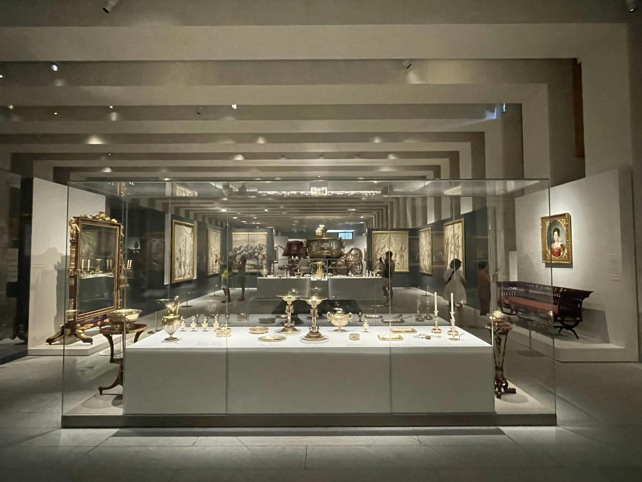 博物馆展览以玻璃箱展示金银文物，墙上有框架艺术品，游客四处走动。