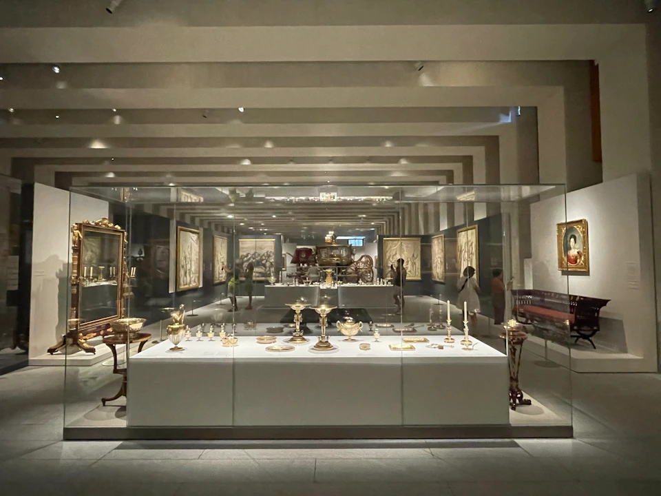 Museum exhibit showcasing gold and silver artifacts in glass cases, with framed artwork on walls and visitors walking around.