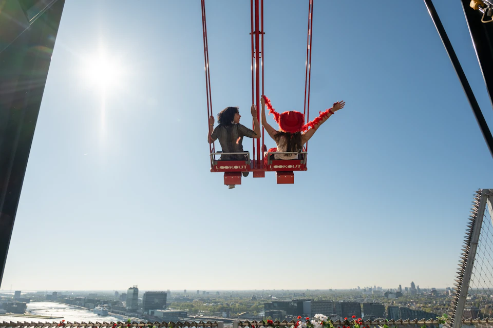 Two people on a red swing ride suspended high above a cityscape under a clear blue sky.