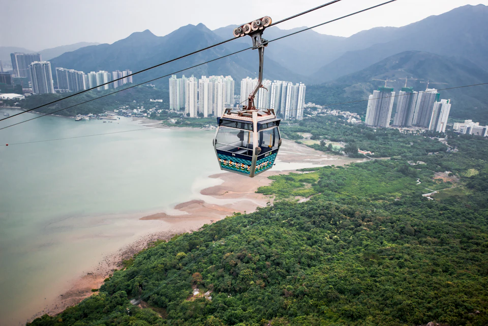 A cable car suspended over lush green hills and a waterfront, with skyscrapers and mountains in the background.