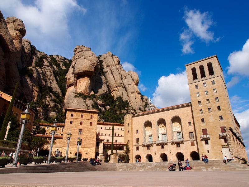 Les bâtiments et monuments historiques en pierre se détachent sur un fond de grandes falaises escarpées, sous un ciel d'un bleu éclatant avec des nuages épars.