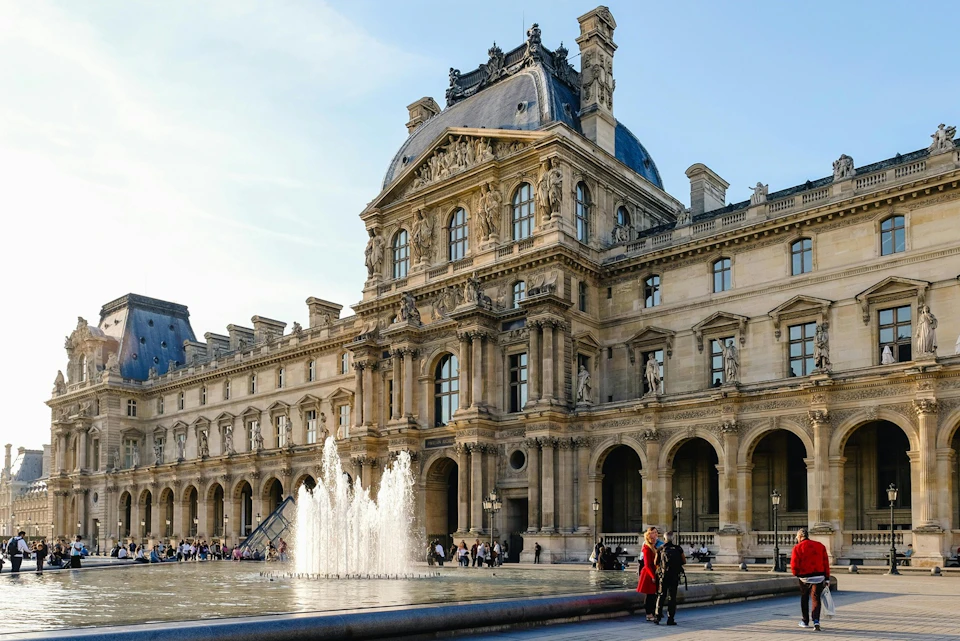A historic building with ornate architecture, a fountain in front, and people walking nearby on a sunny day.