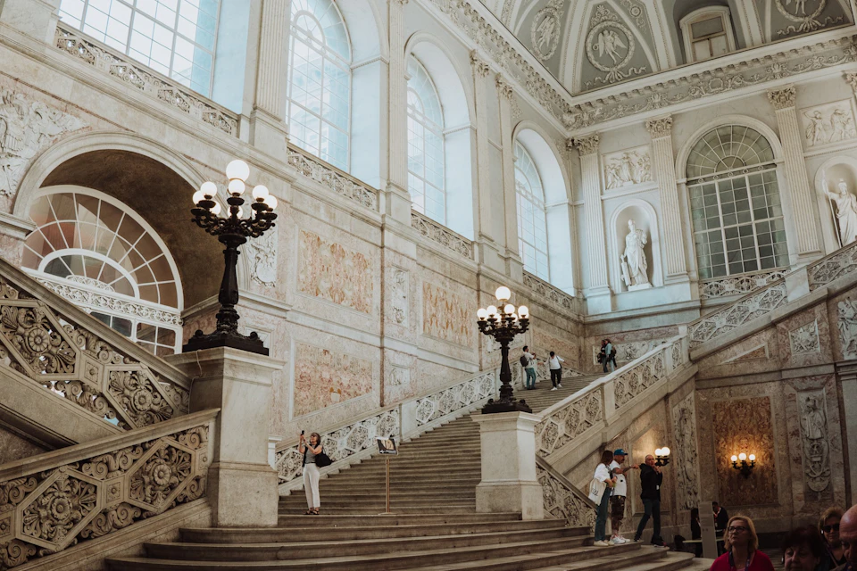 A grand staircase in an ornate building with tall windows, decorative lamps, statues, and several people walking or standing.