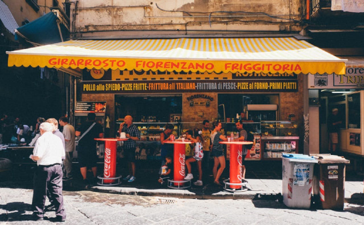 Lidé se shromažďují kolem červených stolů Coca-Cola před stánkem s jídlem se žlutě pruhovanou markýzou a italským nápisem.