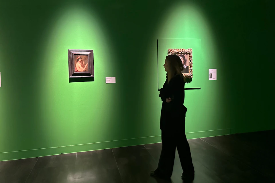 A person walks past paintings displayed on a brightly lit green wall in an art gallery.