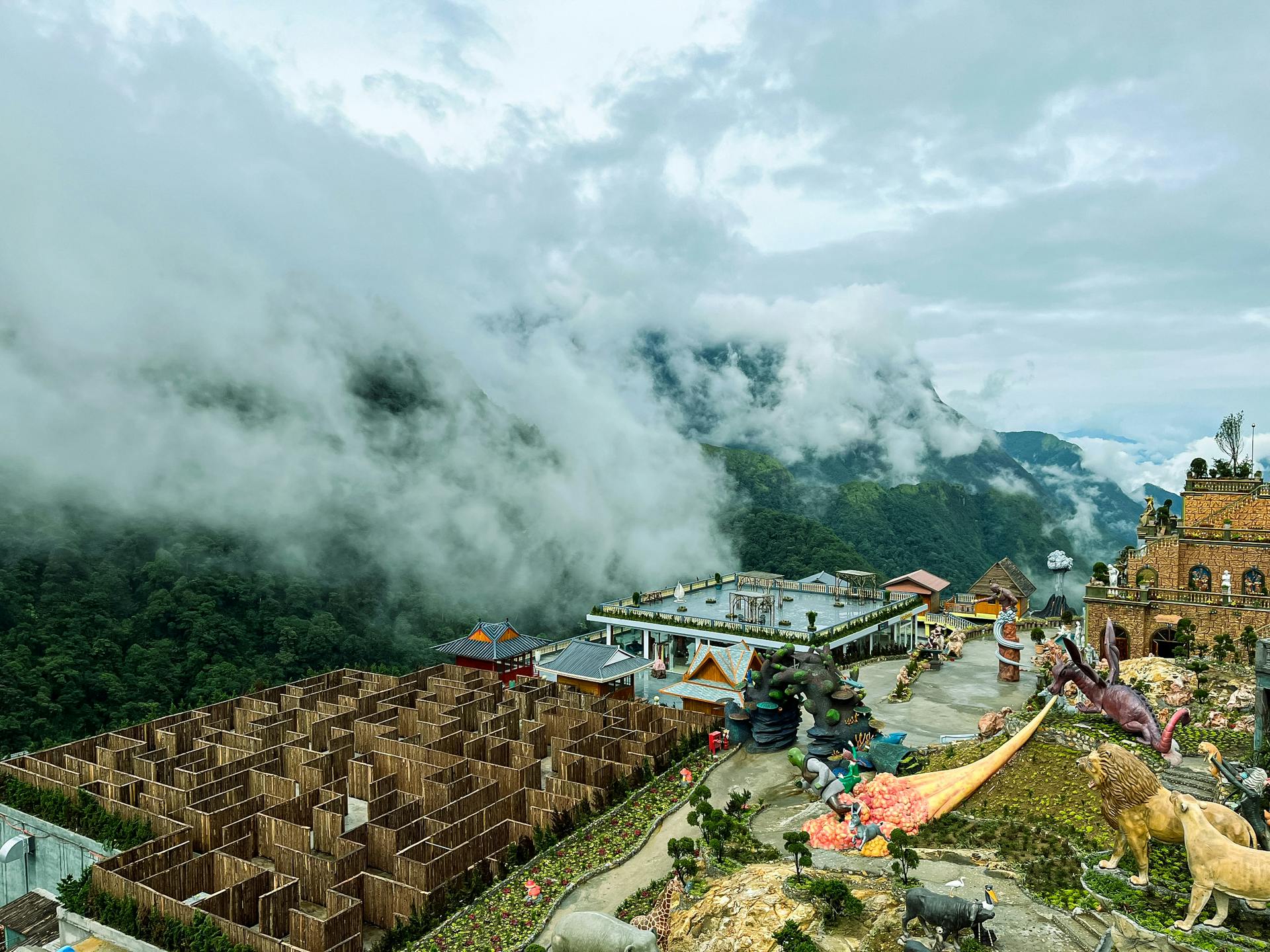 A scenic view of a mountain resort featuring a wooden maze, colorful structures, and surrounding greenery and clouds.