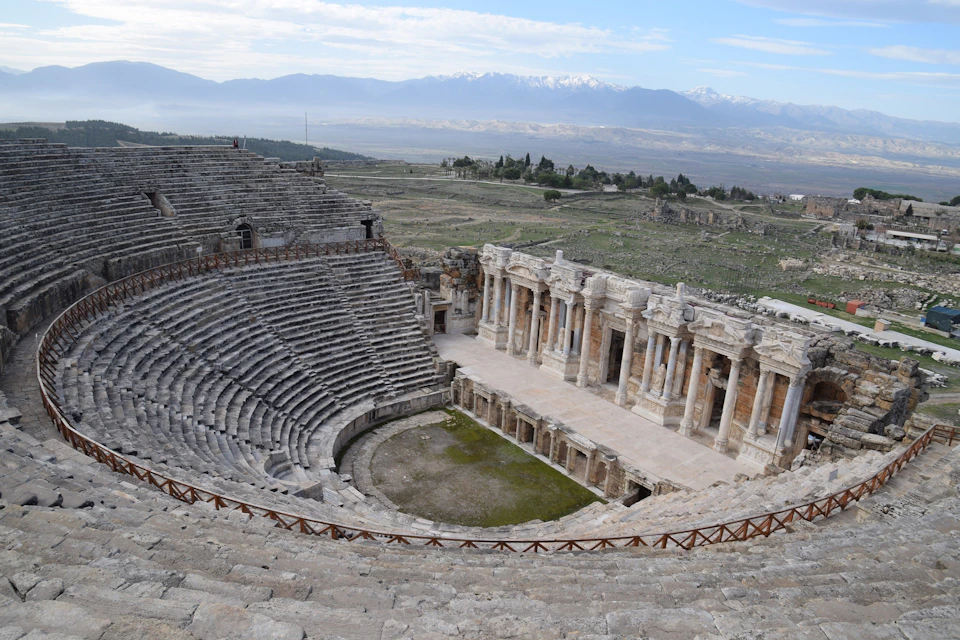 Ancient stone amphitheater with tiered seating and a stage, set in a vast landscape with mountains in the distance.