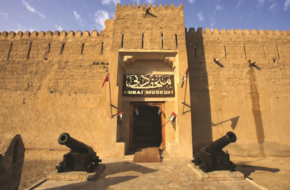 Entrance of the Dubai Museum, featuring an arched doorway flanked by two cannons and flags, set in a historic fortress wall.