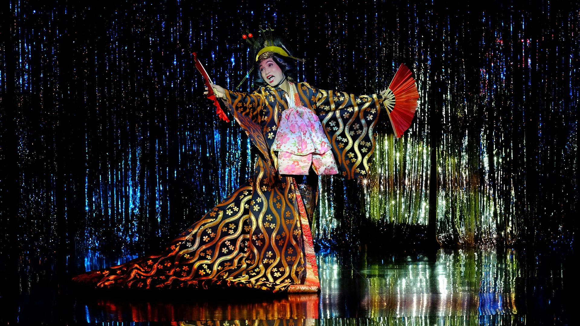 Účinkující v tradičním kostýmu a make-upu držící červené fanoušky na třpytivém modrém pódiu s nataženýma rukama.