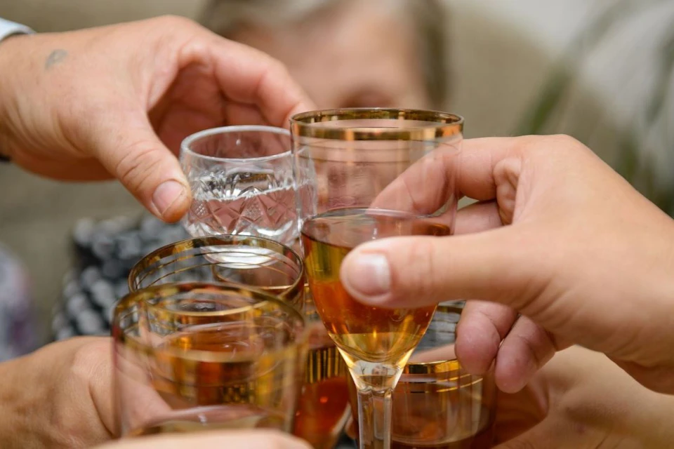 Hands holding glasses of amber liquid clinking together in a toast, with a blurred background.