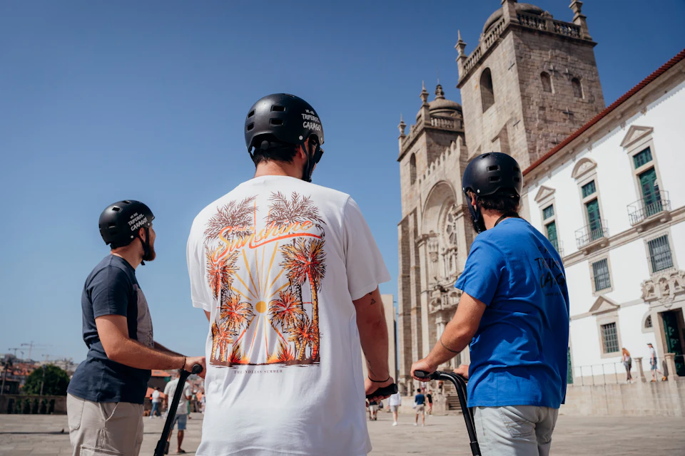Three helmeted individuals on scooters near a historic stone building under a clear blue sky.