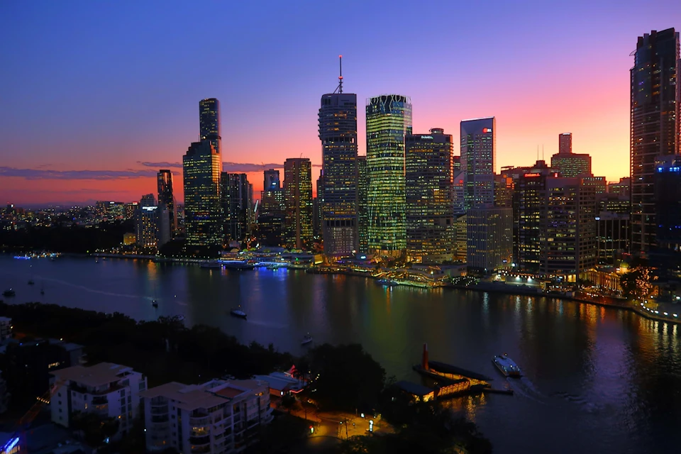A city skyline at sunset, with illuminated skyscrapers by a river. Boats are visible on the water, and trees and buildings line the shore.