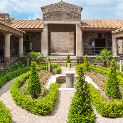 Forntida romersk trädgård med välskötta häckar, små träd, en central fontän, pelare och besökare som utforskar platsen.