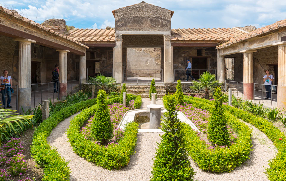 Ancient Roman garden with manicured hedges, small trees, a central fountain, columns, and visitors exploring the site.