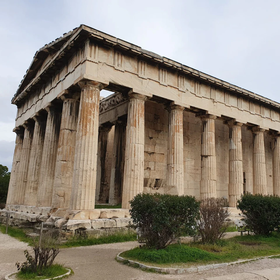 Ancient Greek temple with tall columns, worn and weathered, surrounded by bushes and a footpath under a cloudy sky.