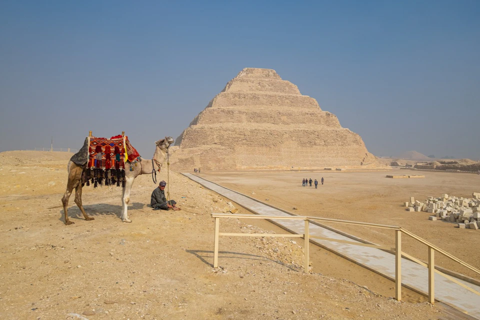 A man sits next to a camel decorated with colorful fabrics in front of a step pyramid with a clear, blue sky overhead.