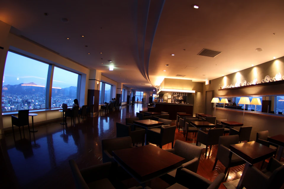 A dimly lit lounge with tables and chairs, large windows showing a cityscape, and a bar area with overhead lighting.