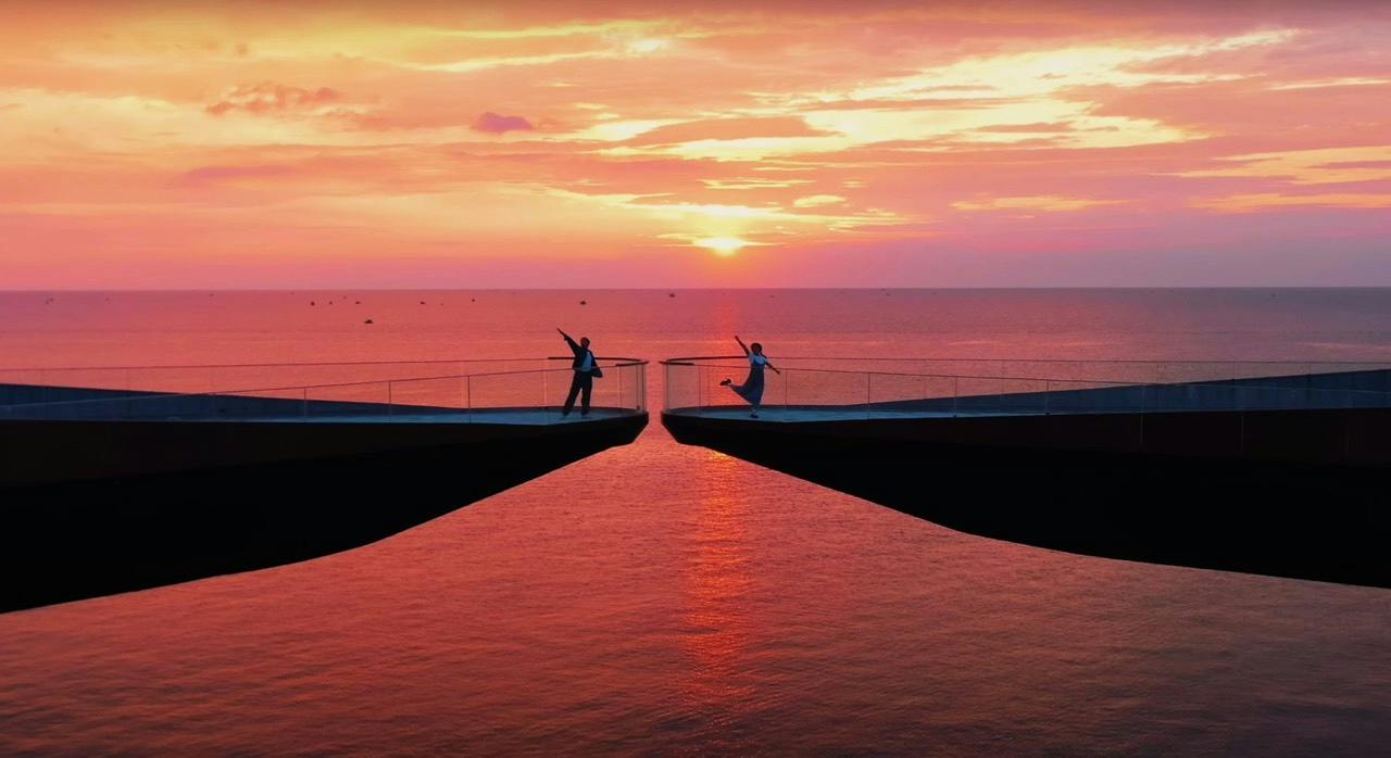 Two people on opposite platforms over water at sunset, with a vibrant orange and pink sky reflected on the water below.