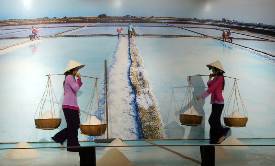 Two people wearing conical hats carry baskets of salt using poles near a mural depicting salt fields.