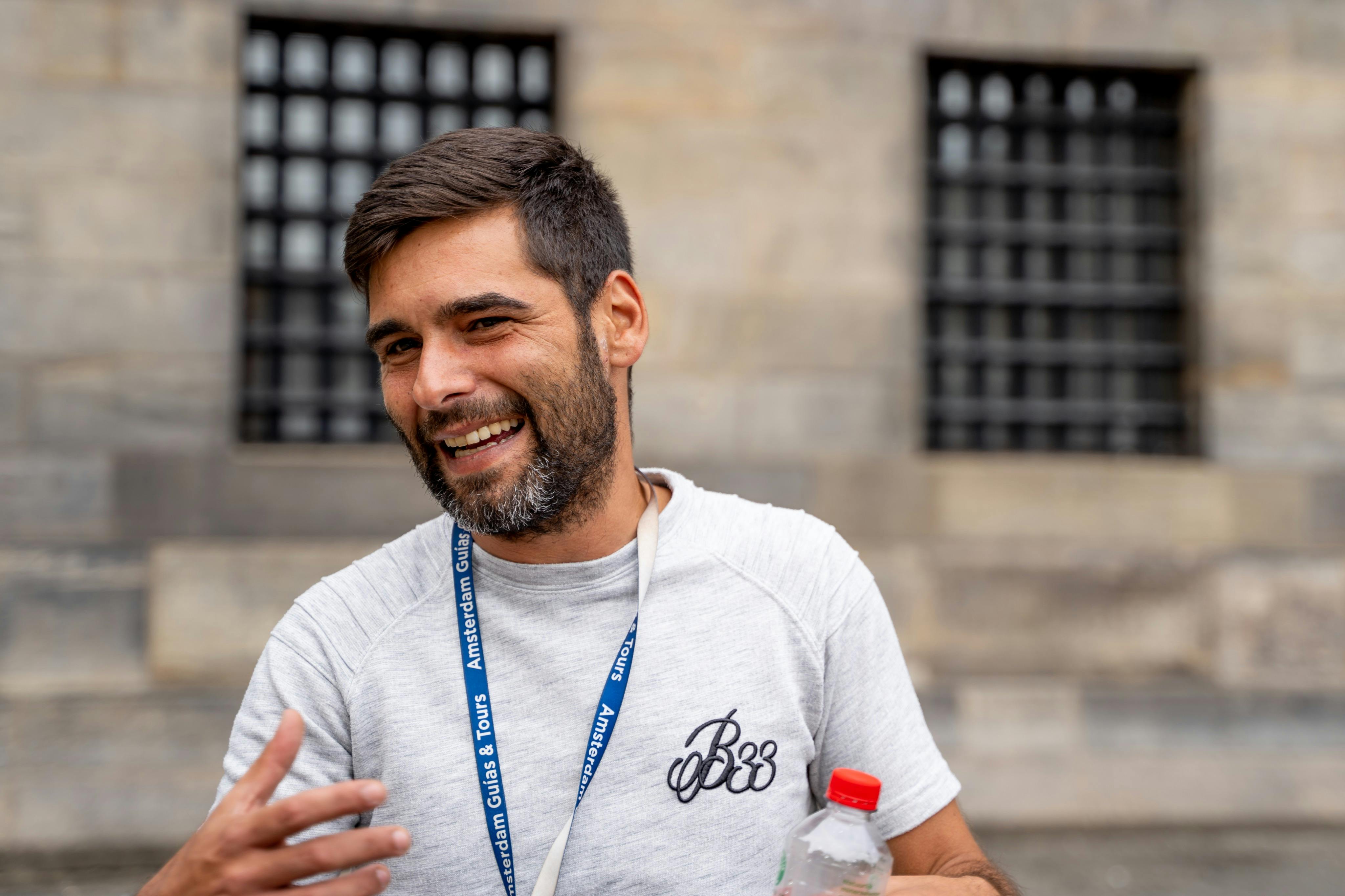 Muž s vousy a krátkými vlasy se usmívá a drží v ruce láhev na vodu. Má na sobě šňůrku a stojí před kamennou budovou.