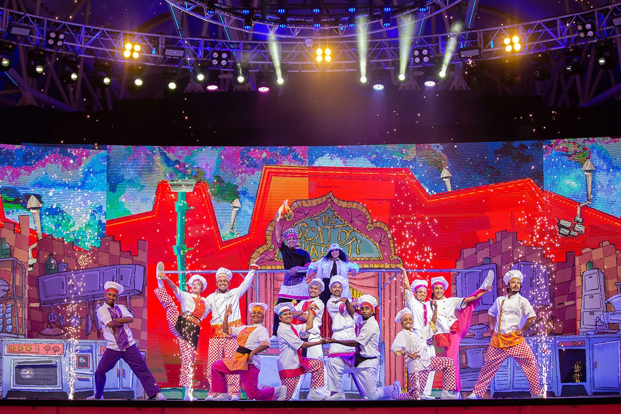 Účinkující v oblečení šéfkuchaře tančí na jasně osvětleném pódiu s barevným, animovaným kuchyňským pozadím.