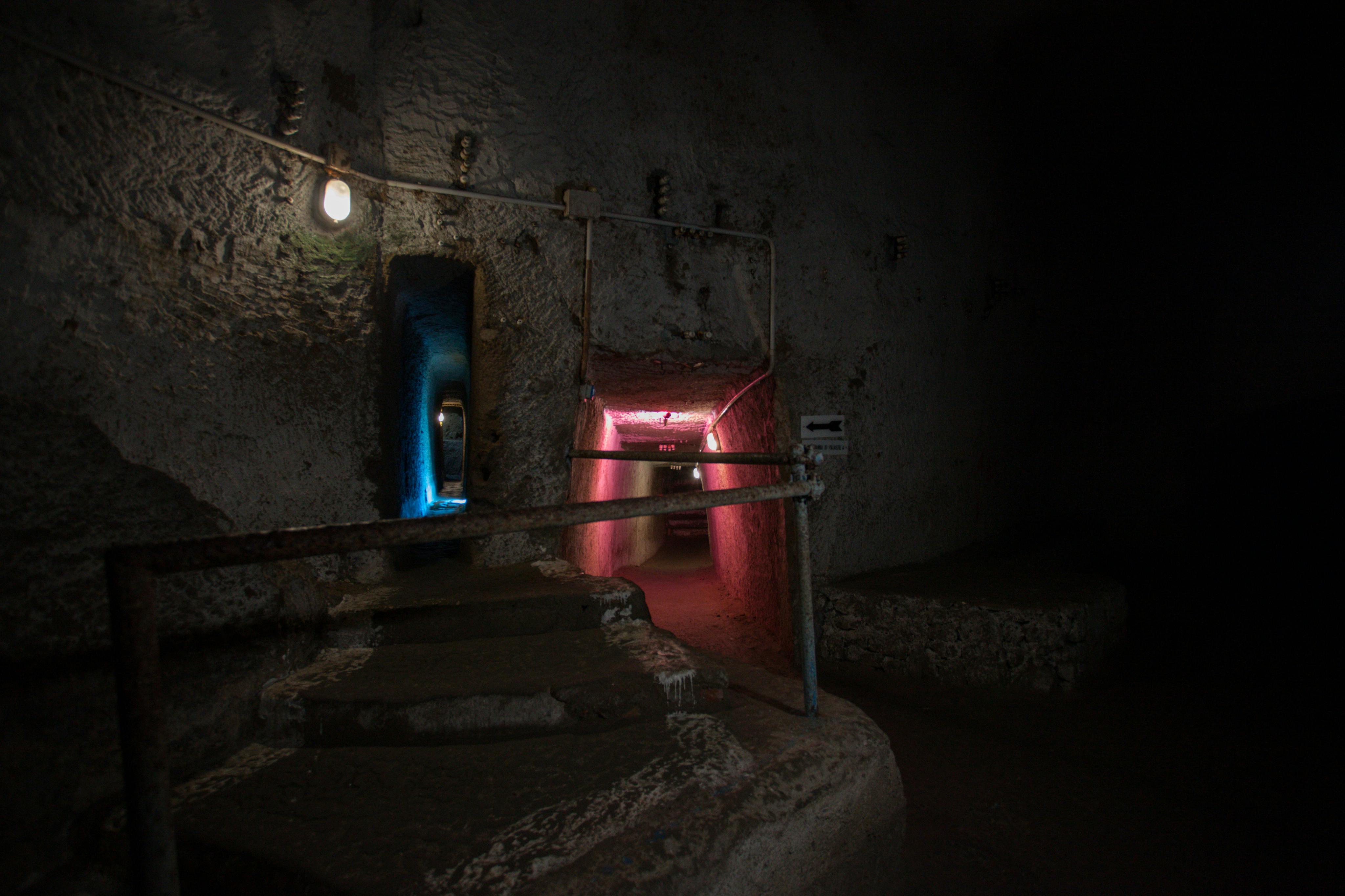 En svagt oplyst underjordisk tunnel med farverige oplyste passager, en glødende lyserød og en anden blå og ru stenvægge.