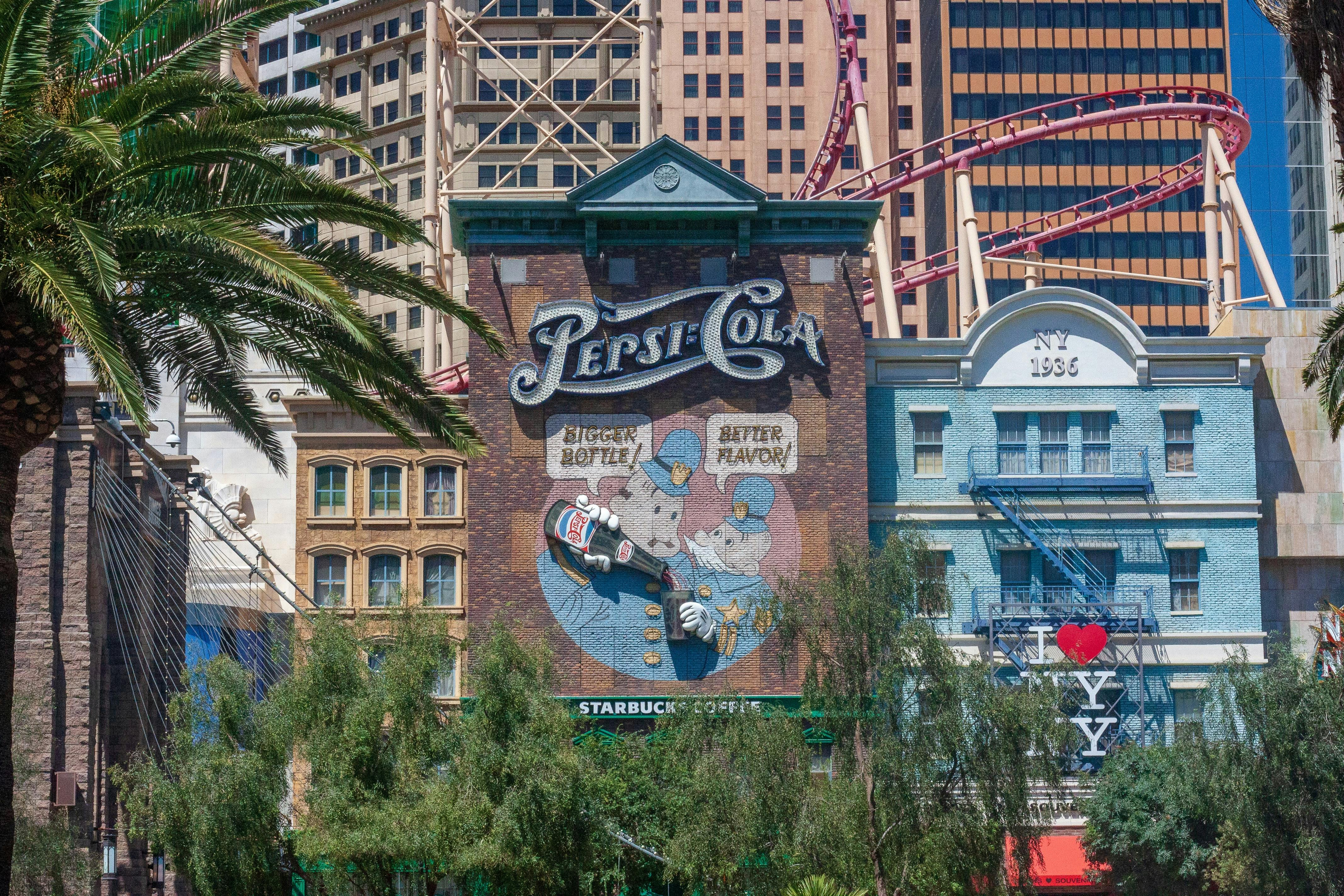 Vintage Pepsi-Cola mural on a city building façade, featuring a waiter serving a bottle. Palms, rollercoaster, and skyscrapers in background.