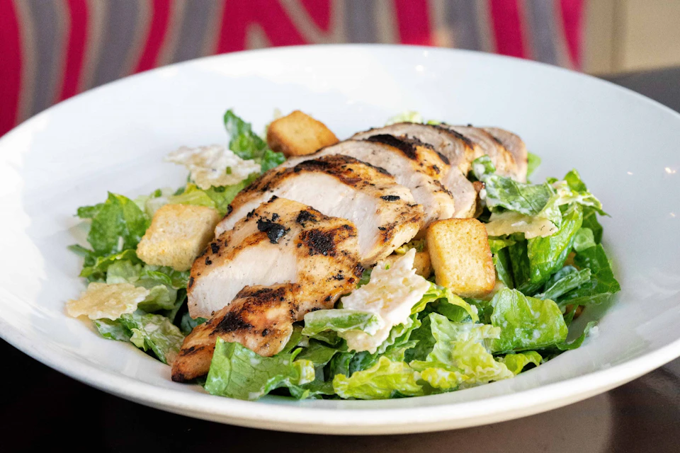A grilled chicken breast sliced over a Caesar salad with romaine lettuce, croutons, and parmesan cheese on a white plate.