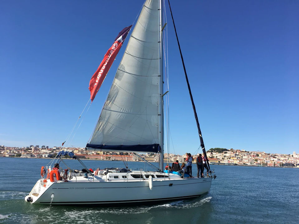A sailboat with a white sail on water, several people on board, and a coastline cityscape in the background under a clear blue sky.