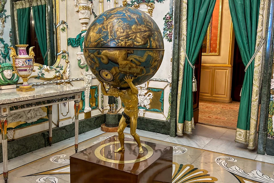 A decorative globe supported by a golden figure in a lavishly decorated room with ornate furniture and green curtains.