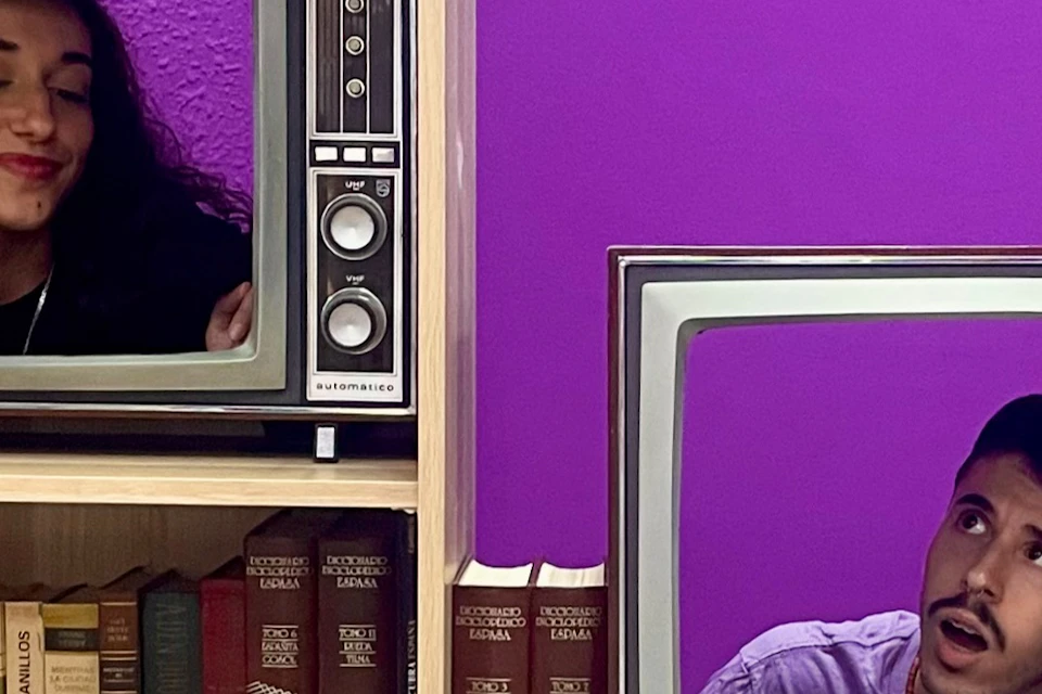 A man and a woman appear on separate vintage TVs placed on a wooden shelf with books and decorative items against a purple wall.
