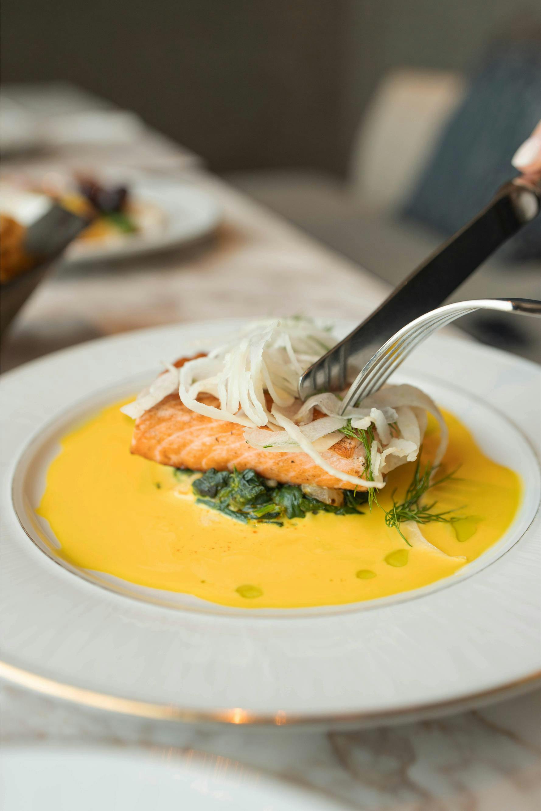 A plated dish of cooked salmon on a bed of green vegetables, topped with shredded garnish, surrounded by a yellow sauce.