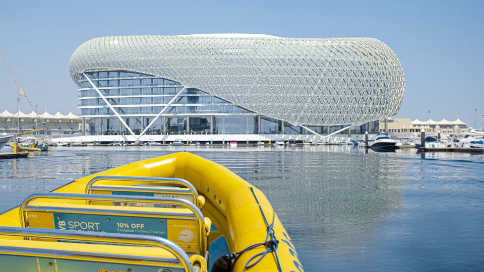 Yellow boat on water faces a modern building with a lattice structure. Floating docks with boats are visible in the background.