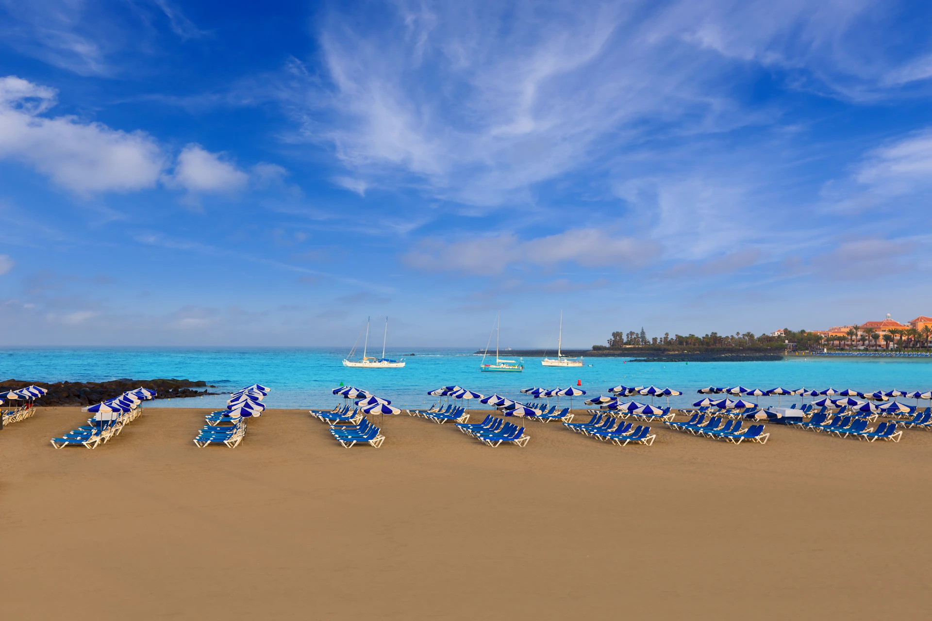 Rows of blue-and-white umbrellas and lounge chairs on an empty beach with sailboats on a calm sea under a clear blue sky.