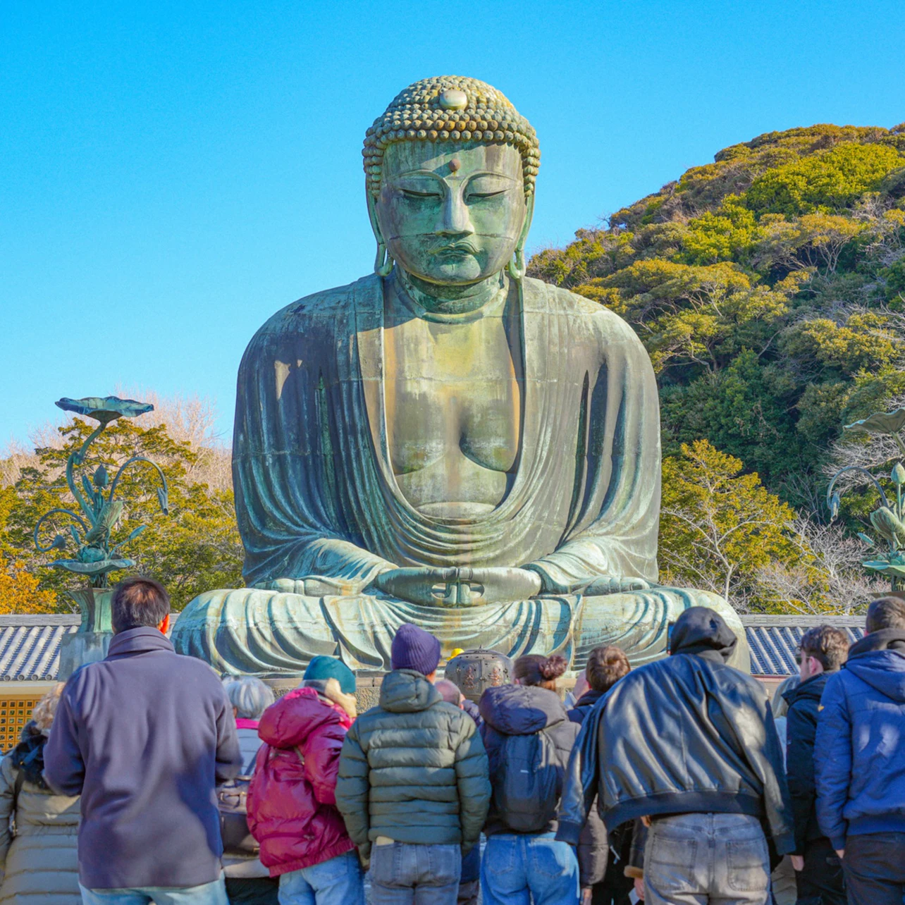 A large Buddha statue outdoors with people gathered in front of it, surrounded by greenery under a clear blue sky.